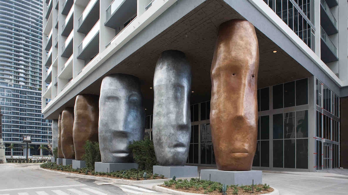 The exterior of the Icon Brickell building showcases five large, abstract sculptures resembling human faces. These sculptures are crafted from materials with varied finishes, presenting a blend of textures. Surrounding landscaping includes low shrubs, and the entrance is framed by expansive glass doors.