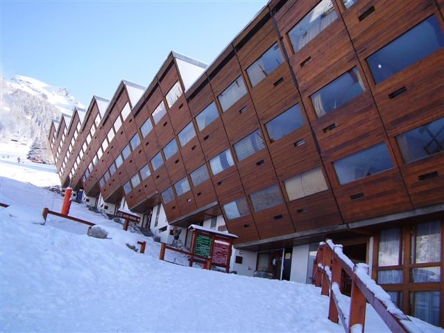 The building features a unique architectural design with slanted wooden panels, blending harmoniously with the snowy mountain landscape. Large windows allow natural light to fill the interior, while a clear sky and snow-covered ground enhance the winter atmosphere.