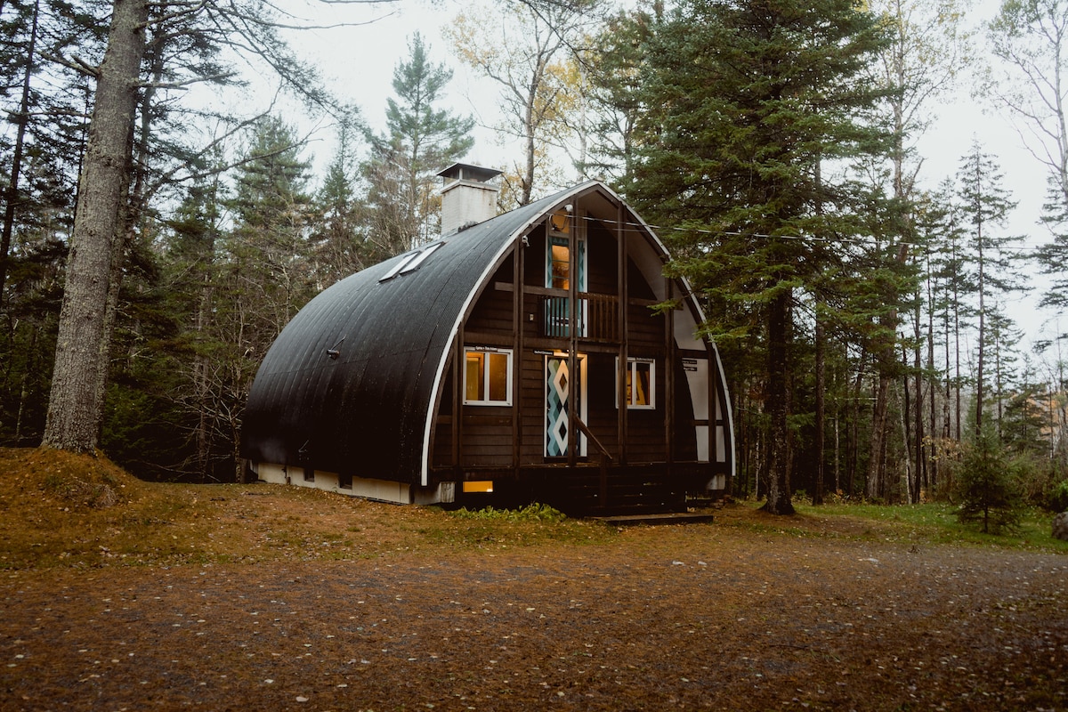 The cabin's exterior showcases a unique A-frame design with a curved roof and wooden siding. Large windows allow natural light to enter, while the surrounding forest adds a serene backdrop. A gravel path leads to the entrance, framed by autumn foliage.