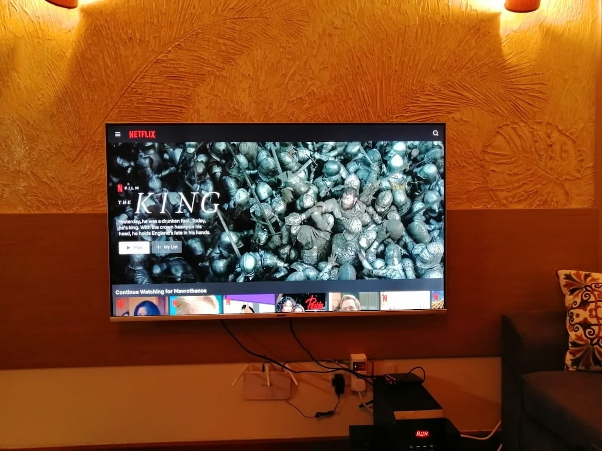 A flat-screen television is mounted on a textured wall, displaying the Netflix homepage. Surrounding lighting enhances the ambiance. A media console is positioned below, featuring various electronic devices and cables. A cozy seating area is visible on the side, decorated with patterned cushions.