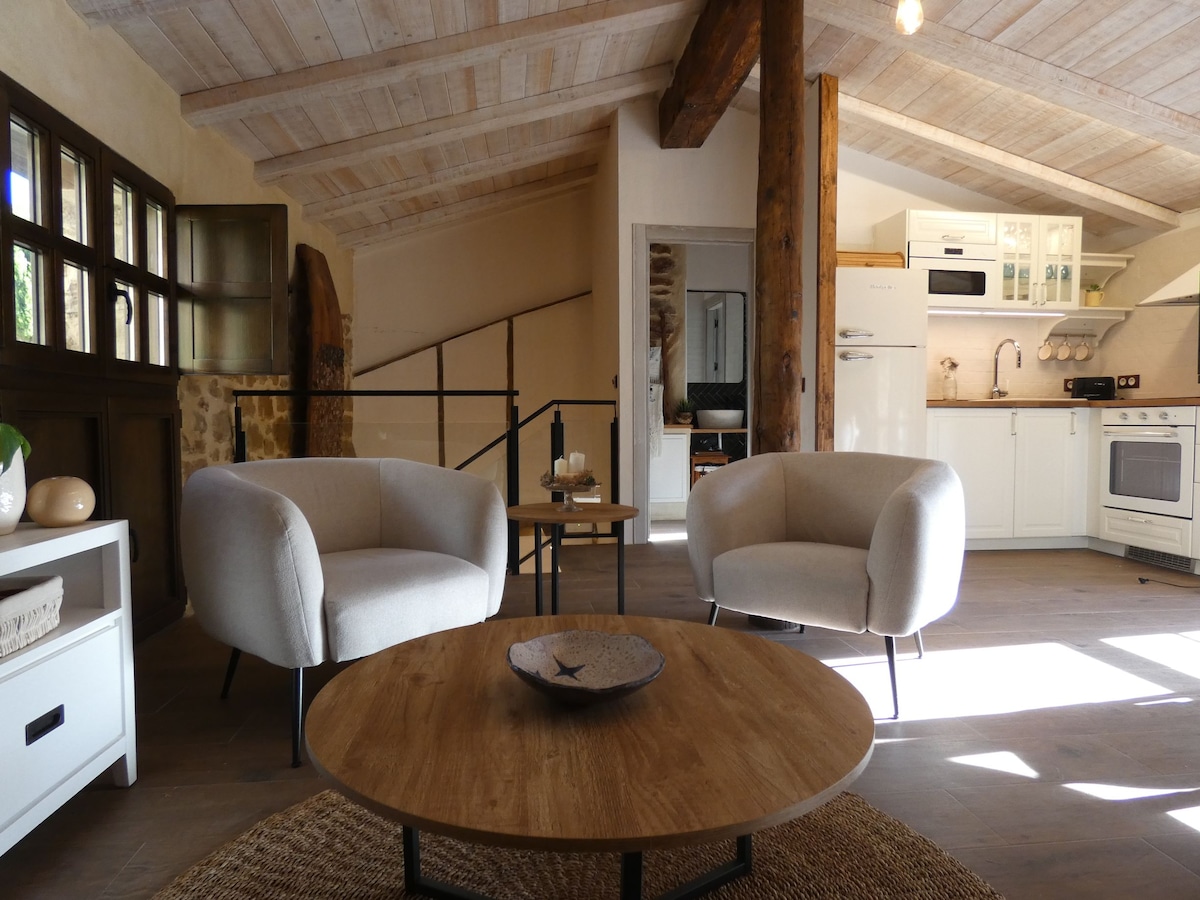 A spacious living area features two comfortable armchairs facing a wooden coffee table. Natural light fills the room through large windows, highlighting the rustic wooden beams and open kitchen in the background. The decor integrates natural textures, creating a welcoming ambiance.