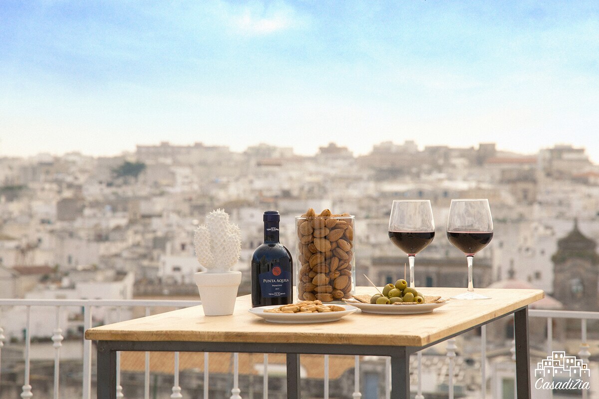 A wooden table is set on a terrace with a panoramic view of the white city. Two glasses of red wine accompany a bottle of local wine and a jar filled with almonds. A plate of cookies and a small bowl of olives add to the inviting display.