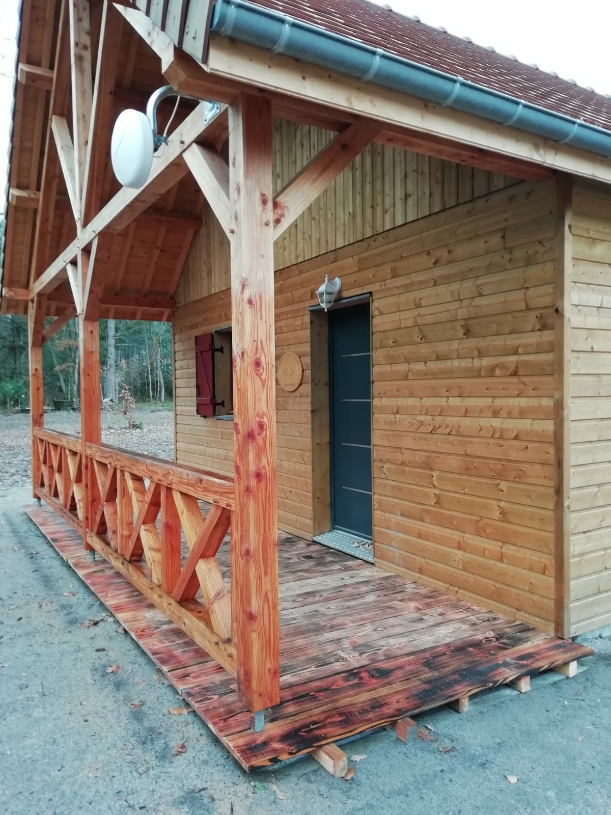 A wooden chalet exterior showcases a large covered porch with a railing made of sturdy logs. The door is framed by warm-colored wooden siding, and a decorative round element adds a rustic touch. Natural surroundings are visible, enhancing the serene setting.