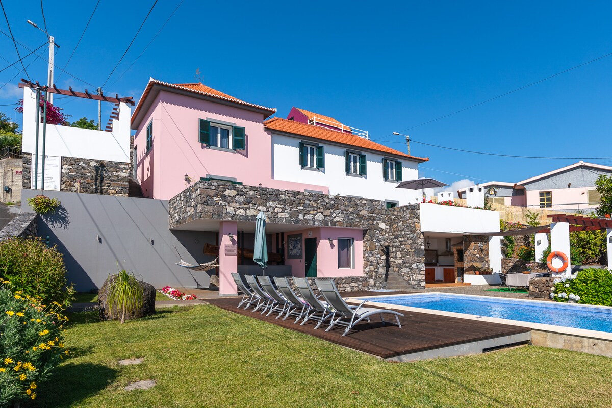 A two-story villa is showcased, featuring stone facade accents and a bright pink upper level. A spacious swimming pool is framed by sun loungers on a wooden deck, surrounded by lush greenery and vibrant flowers. Clear blue skies provide a serene backdrop.