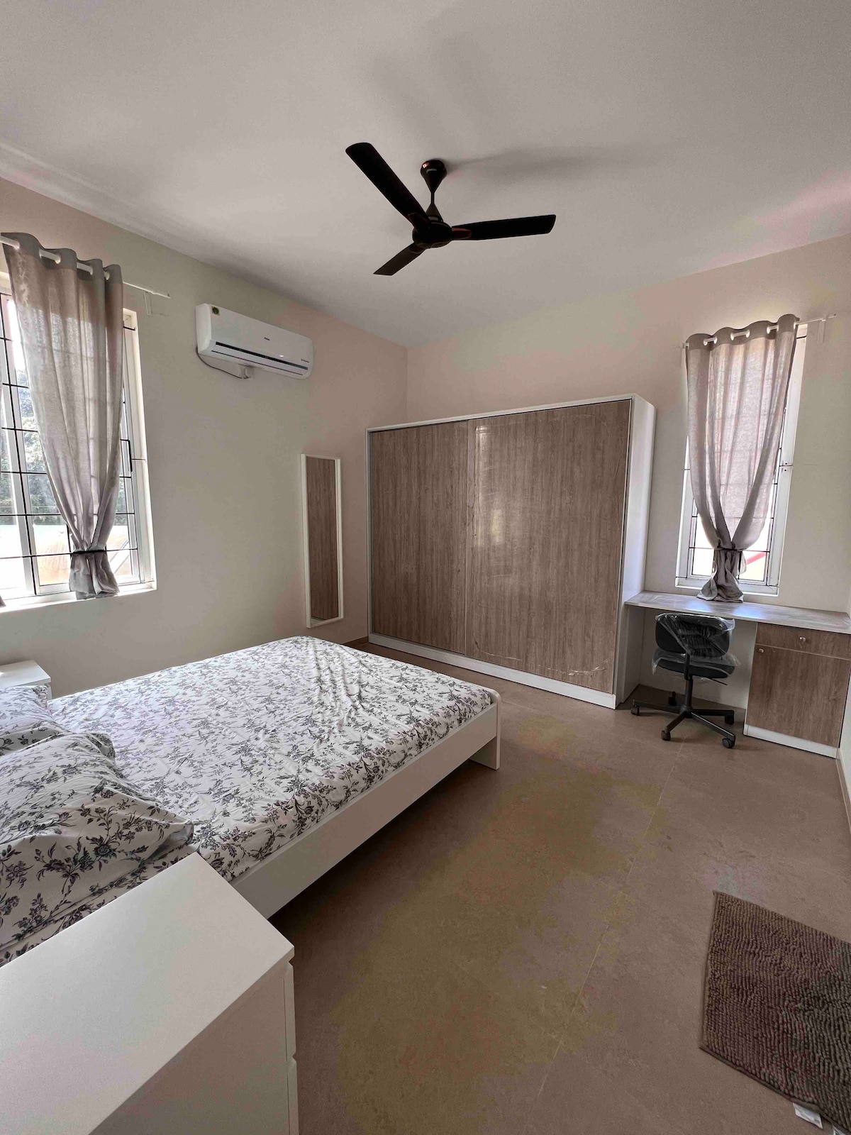 A cozy bedroom features a spacious bed with floral bedding. Natural light enters through two windows adorned with light curtains. A sleek wooden wardrobe stands beside the bed, and a workspace with a chair is positioned against the wall, creating a functional living area.