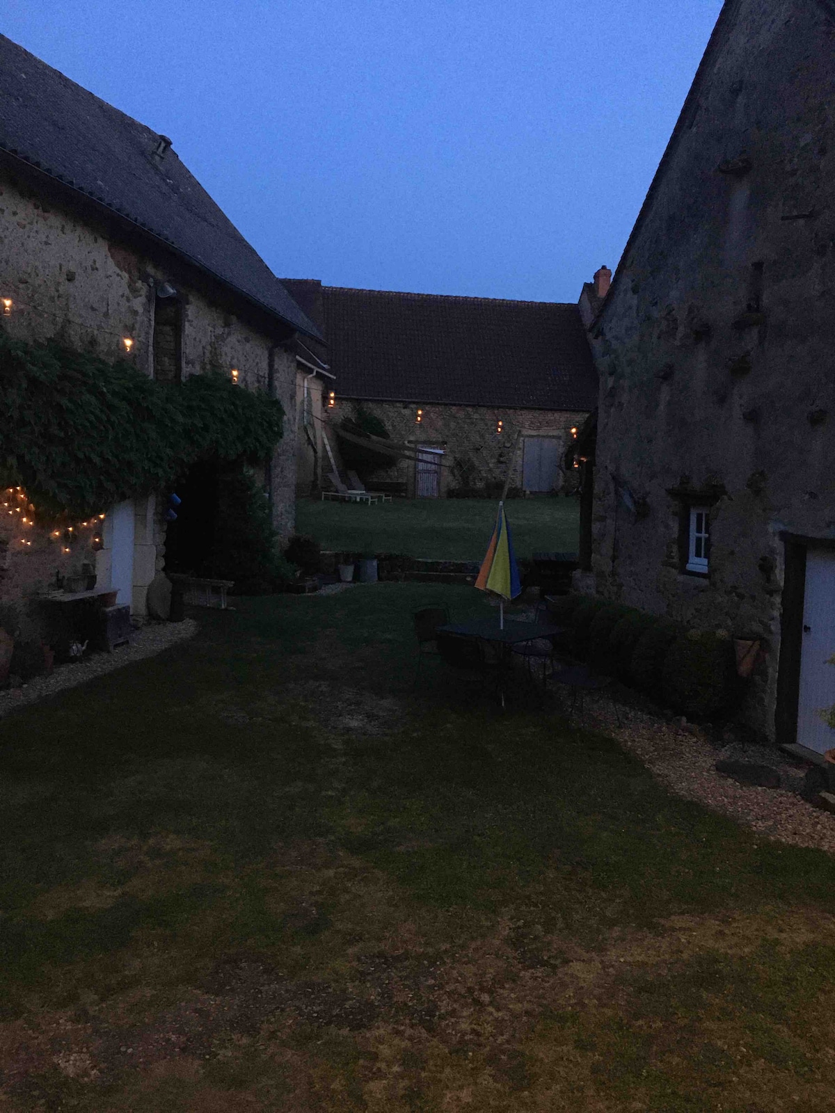 The exterior area is set during twilight, revealing a grassy courtyard surrounded by rustic stone walls. Soft lights illuminate the space, reflecting a calm atmosphere. A patio area with tables and chairs, along with a decorative flag, can be seen, contributing to the overall serene environment.