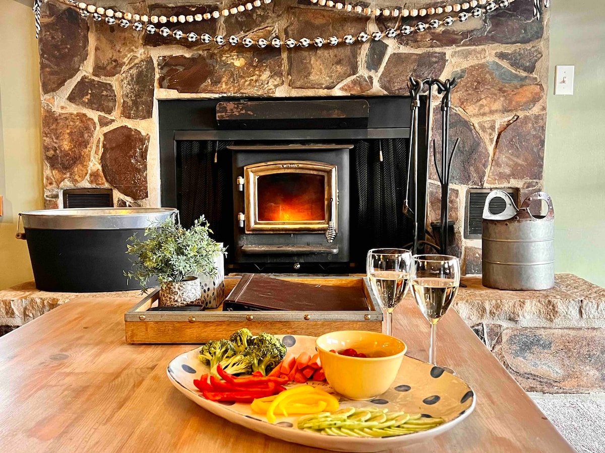 A stone fireplace serves as a focal point, with a warm fire visible inside. A wooden table displays a colorful plate of assorted vegetables and dipping sauce, accompanied by two elegant glasses. Decorative elements enhance the cozy ambiance.