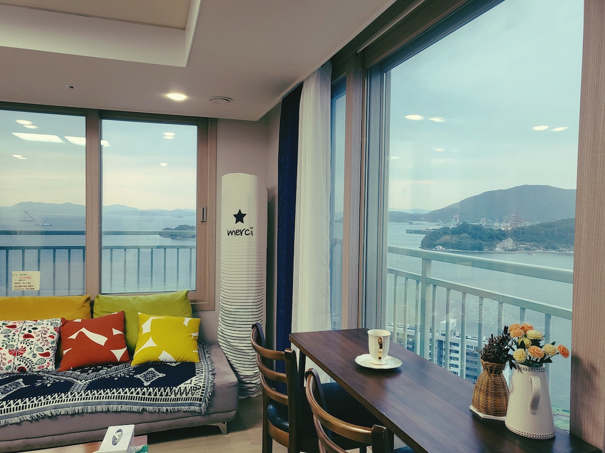 A seating area is positioned near large windows, offering panoramic views of the sea and nearby islands. The space features a sofa adorned with colorful cushions and a wooden dining table with decorative flowers. Soft natural light illuminates the room, enhancing the view.