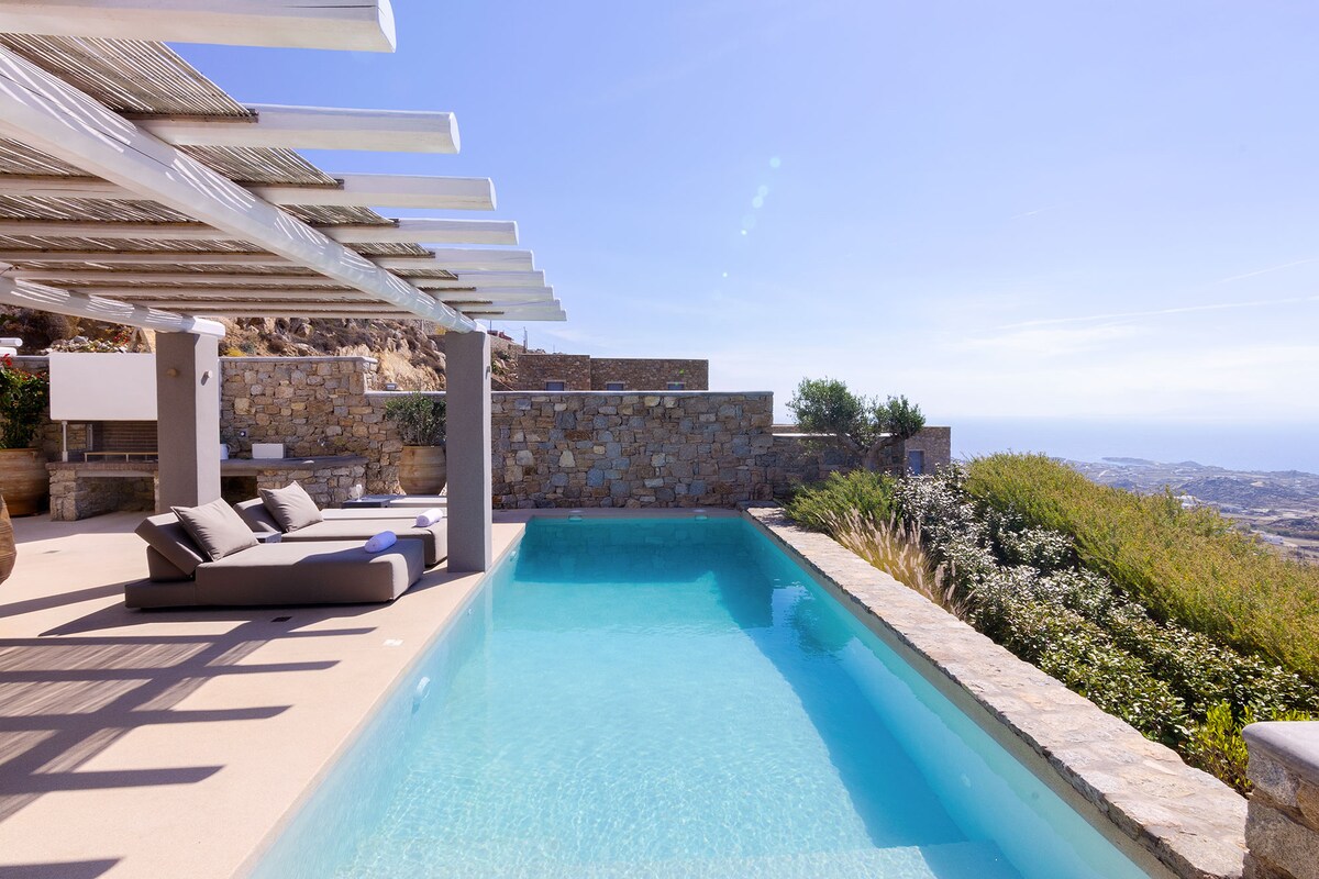 A private swimming pool is positioned under a shaded pergola, surrounded by smooth stone walls and lush greenery. Expansive views of the landscape and sky are visible, enhancing the tranquil outdoor setting. Comfortable loungers provide an area for relaxation beside the pool.