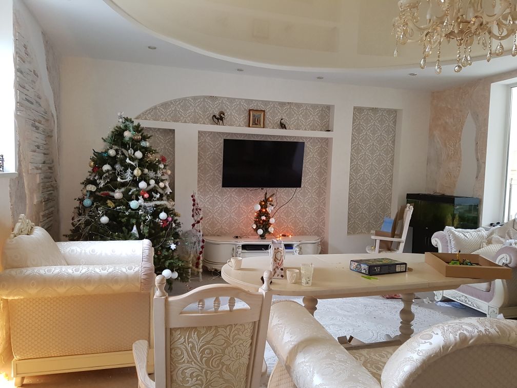 A stylish living area features a Christmas tree adorned with decorations. A large television is mounted on a decorative wall. Comfortable seating is arranged around a coffee table, creating a welcoming atmosphere. A subtle aquatic feature can be seen in the background.