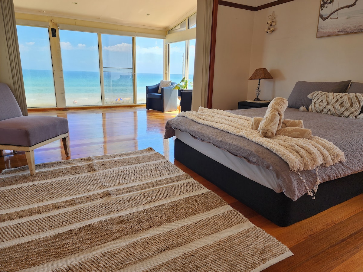 A spacious bedroom features a large, comfortable bed with a textured blanket draped across it. Natural light floods the room through expansive glass doors, which offer stunning views of the beach and ocean. A cozy seating area is visible in the background.