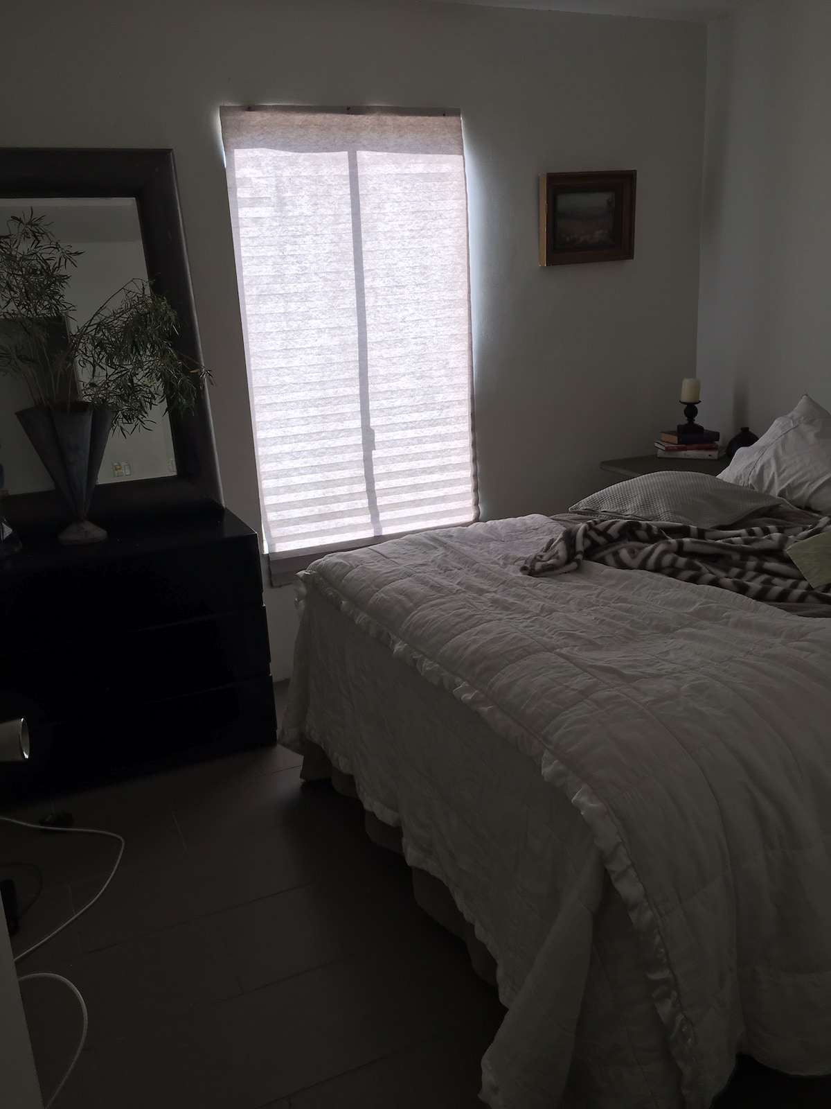 A serene bedroom features a spacious bed covered with a light quilt and layered blankets. Natural light filters through a partially closed window adorned with vertical blinds. A mirror and a decorative vase are positioned on a black dresser, with a small stack of books nearby.