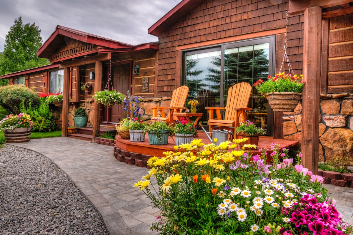 A welcoming entrance features a flower-filled landscape with vibrant blooms. A stone path leads to a wooden porch furnished with two adirondack chairs, creating a lovely space for outdoor relaxation. Window views showcase the garden's lush greenery and colorful hanging planters.