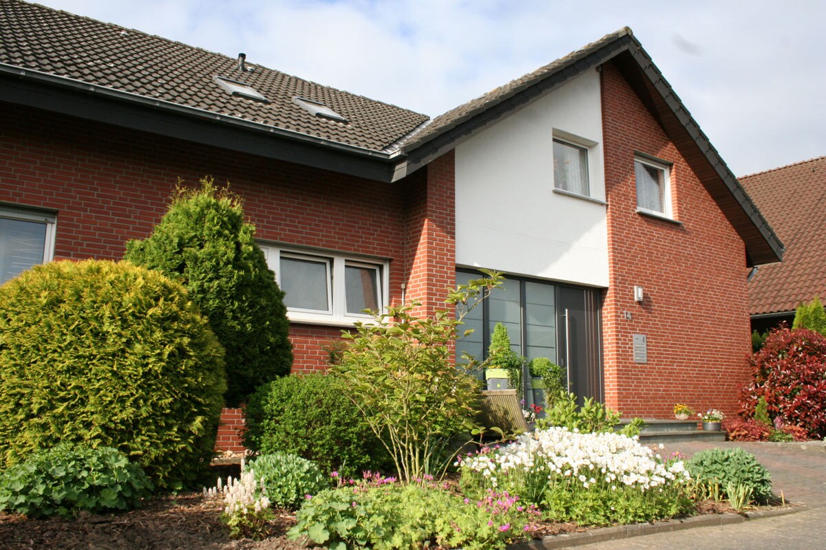 The exterior of a modern brick building is presented, featuring a sloped roof and large windows. Lush greenery, including shrubs and flowerbeds with various blooms, enhances the entrance area. A clear pathway leads to the front door, with additional landscaping visible on either side.