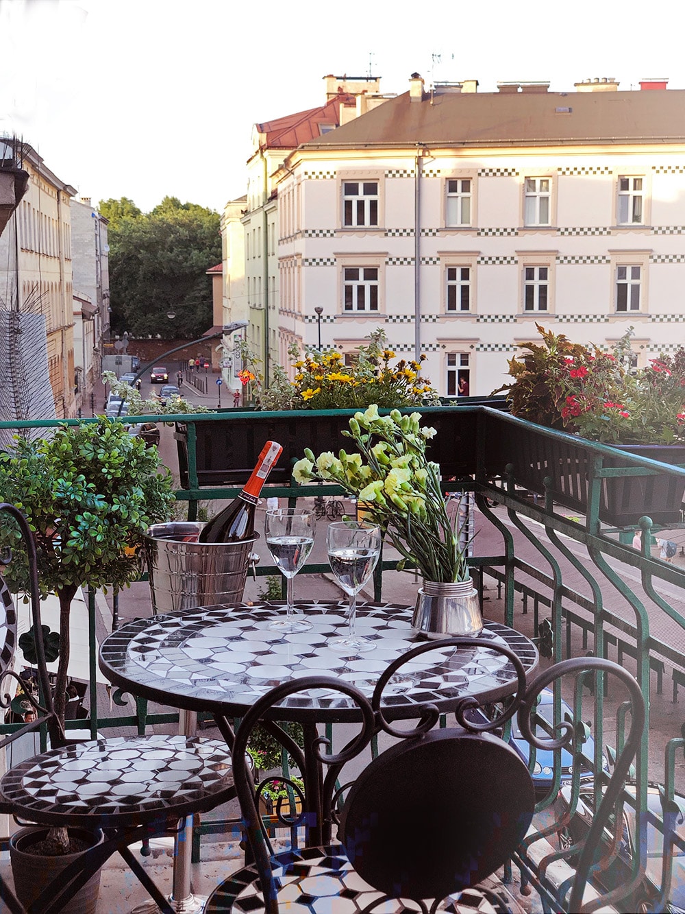 A balcony table with a tiled surface is set with wine glasses and a bottle of champagne, surrounded by potted plants. The view features nearby buildings and street scenes, with colorful flowers adding charm to the setting.