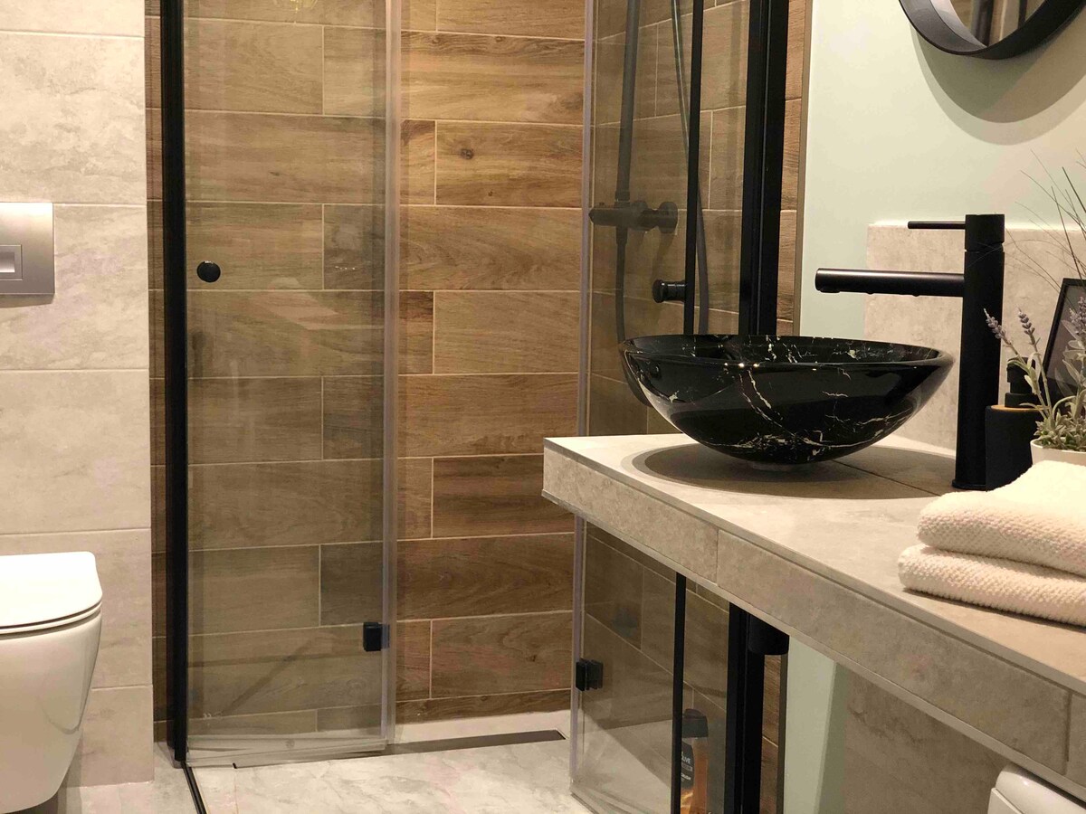 A modern bathroom features a standing shower enclosed in glass, complemented by a stylish black vessel sink and a sleek countertop. Natural wood tones adorn the walls, creating a warm ambiance, while neatly arranged towels and decorative elements add a touch of elegance.