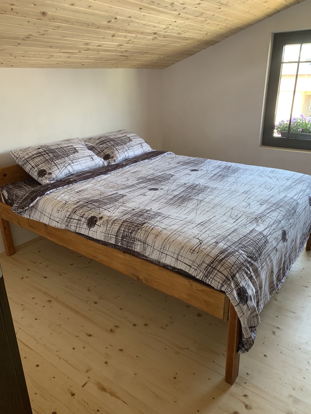 A spacious king bed is positioned against a wall, fitted with a neutral-toned bedspread featuring a subtle pattern. Pillows are arranged neatly at the head of the bed. Natural light enters through a window, which offers a view of the outside greenery.