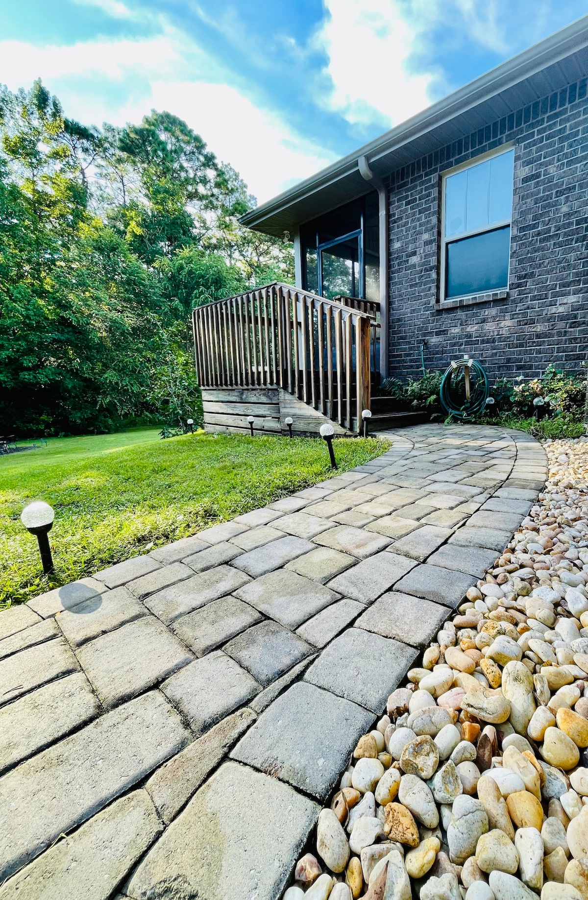 A stone pathway leads to a private entrance, bordered by green grass and decorative pebbles. The area is illuminated by small, low-profile lights. The building's exterior features a mix of brick and large windows, surrounded by trees providing privacy.