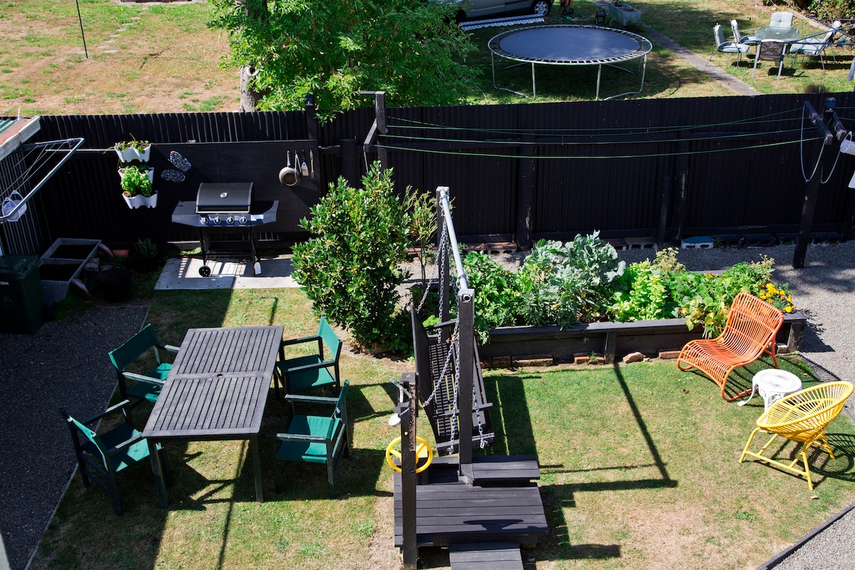 An outdoor area is presented, featuring a combination of seating and play options. A square dining table surrounded by chairs lies on green grass. A wooden swing set and trampoline can be seen in the background, along with a BBQ grill and vegetable garden beds.
