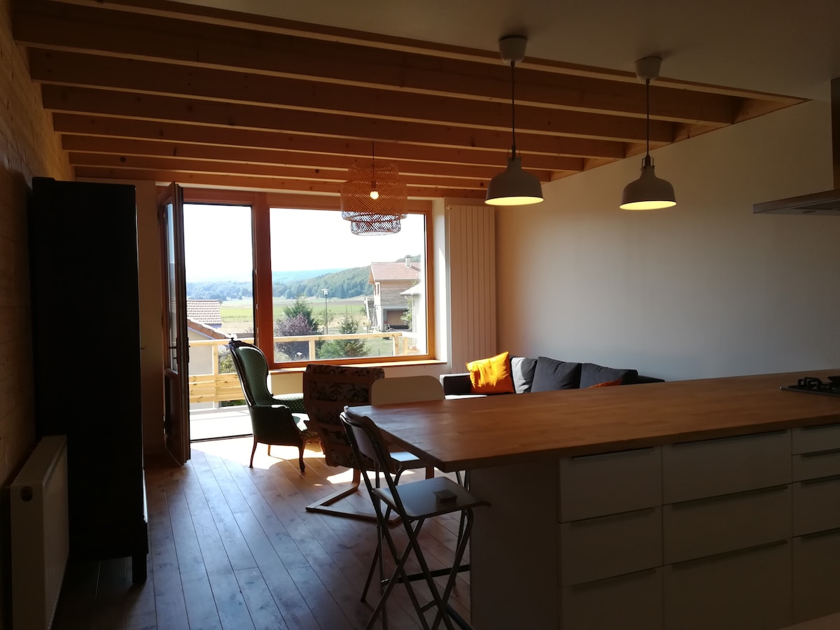 A bright and airy living space features large windows that offer views of the surrounding landscape. Wooden beams accent the ceiling, while warm lighting hangs above a kitchen island. A cozy seating area includes a gray sofa and a colorful accent chair.