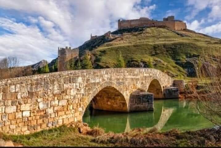 A stone bridge with three arches spans over a tranquil green-tinted waterway, reflecting the nearby landscape. In the background, a historic castle is perched atop a hill, surrounded by rolling fields and a cloudy sky.