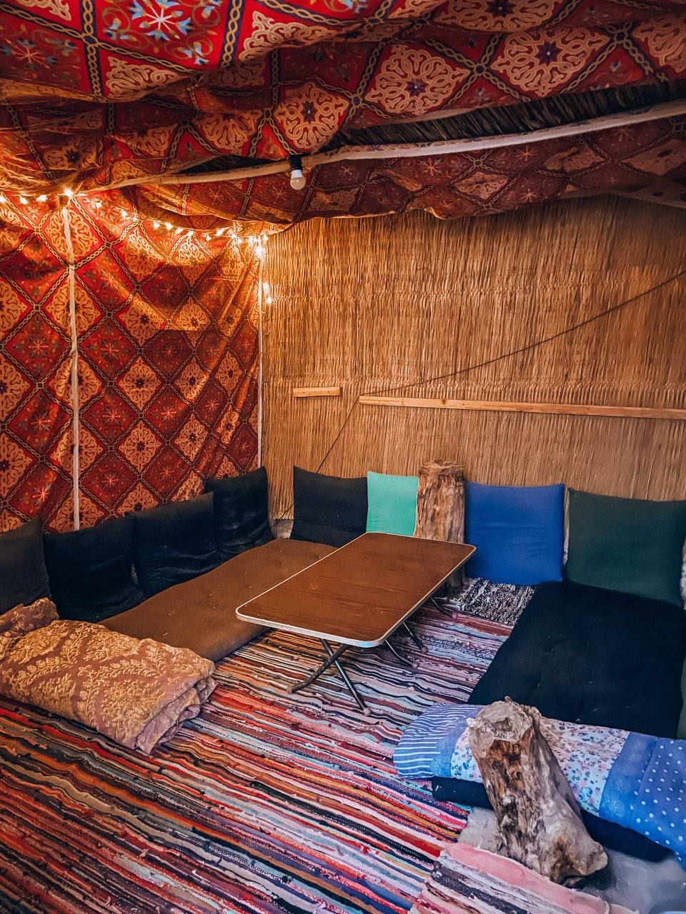 A cozy indoor space features low seating on vibrant patterned rugs, with colorful cushions arranged on the floor. A simple wooden table is positioned at the center, and soft string lights create a warm glow against the richly patterned fabric draping the walls.