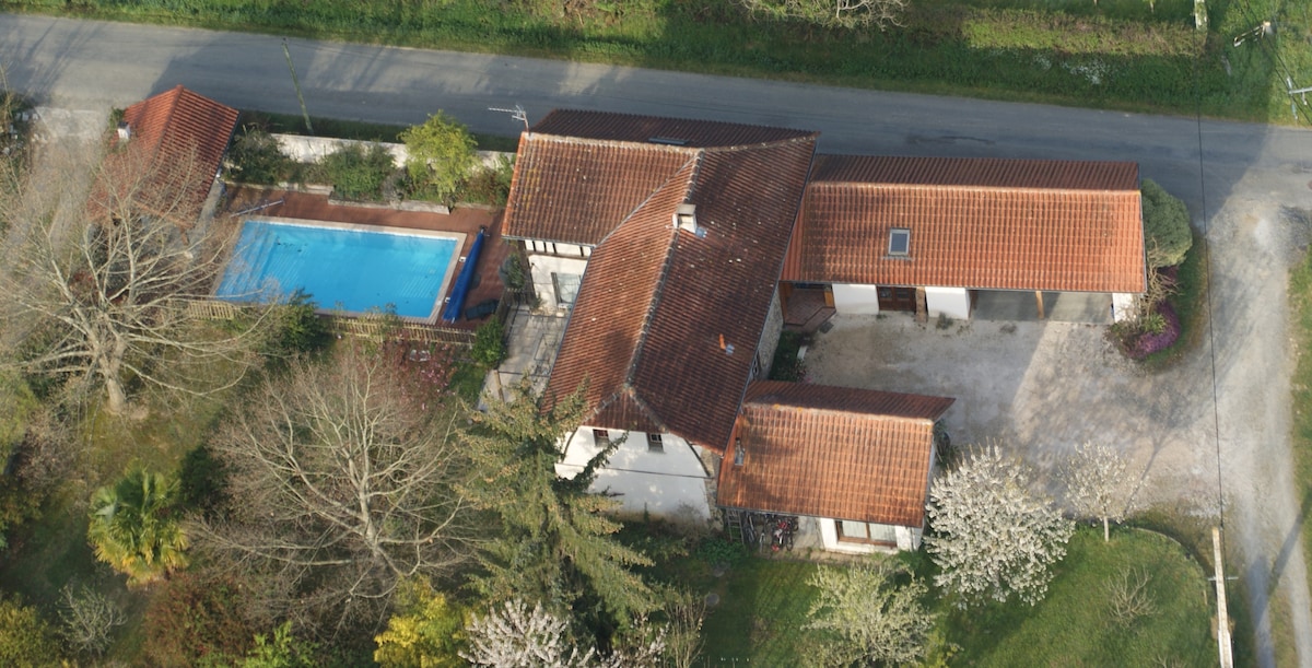 An aerial view of the property showcases a spacious house with a red-tiled roof and a large pool surrounded by greenery. The pool and outdoor spaces are nestled within a tranquil setting, with trees and shrubs enhancing the peaceful environment.