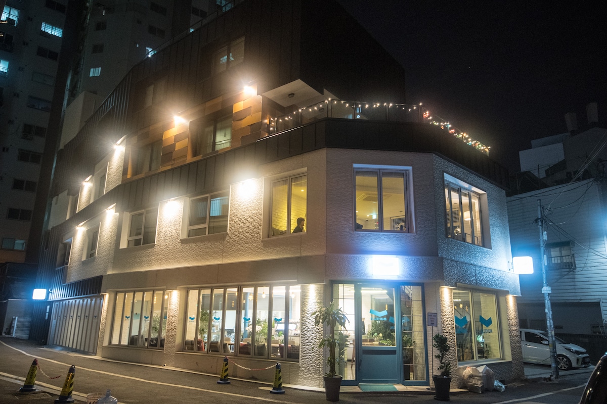 The exterior of the guest house is illuminated at night, showcasing a modern three-story building with large windows and decorative lighting. A cozy ambiance is created by warm lights adorning the upper levels. The surrounding area features a clean, well-maintained environment.