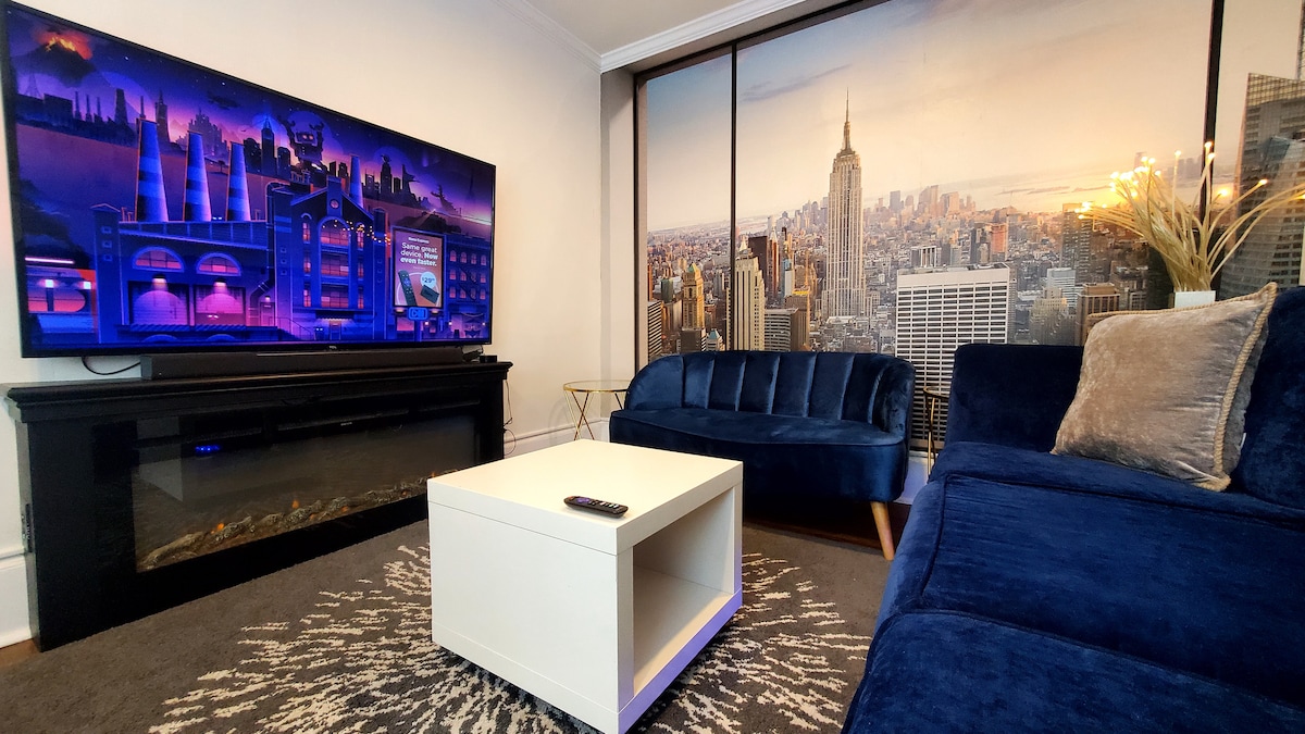 Relax in this modern and elegant space, decorated with a stunning city mural and cozy atmosphere. Enjoy a unique experience with a comfortable couch, electric fireplace, and a big screen for your entertainment moments.