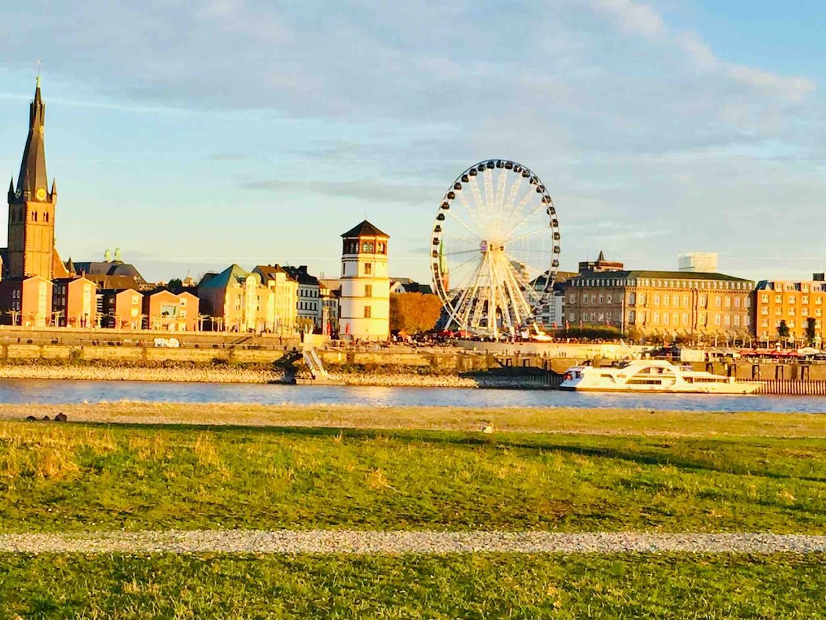 A waterfront view features a historic skyline with a tall church spire to the left and a prominent Ferris wheel in the background. The scene is complemented by a gentle river with a boat and grassy areas in the foreground, all under a clear sky.