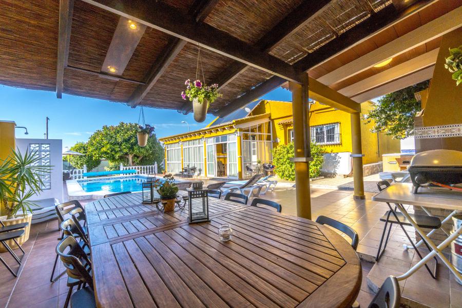 An outdoor dining area is furnished with a large wooden table surrounded by multiple chairs. A shaded roof provides cover. In the background, a pool glistens under clear skies, with loungers positioned nearby. Lush greenery can be seen framing the space.