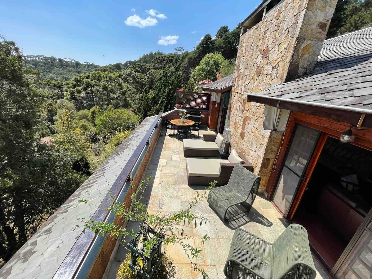 A spacious terrace is seen featuring comfortable lounges and two modern tables. Stone walls blend with the natural surroundings, creating an inviting atmosphere. Green trees and a clear sky enhance the tranquil setting, ideal for outdoor relaxation and gathering.