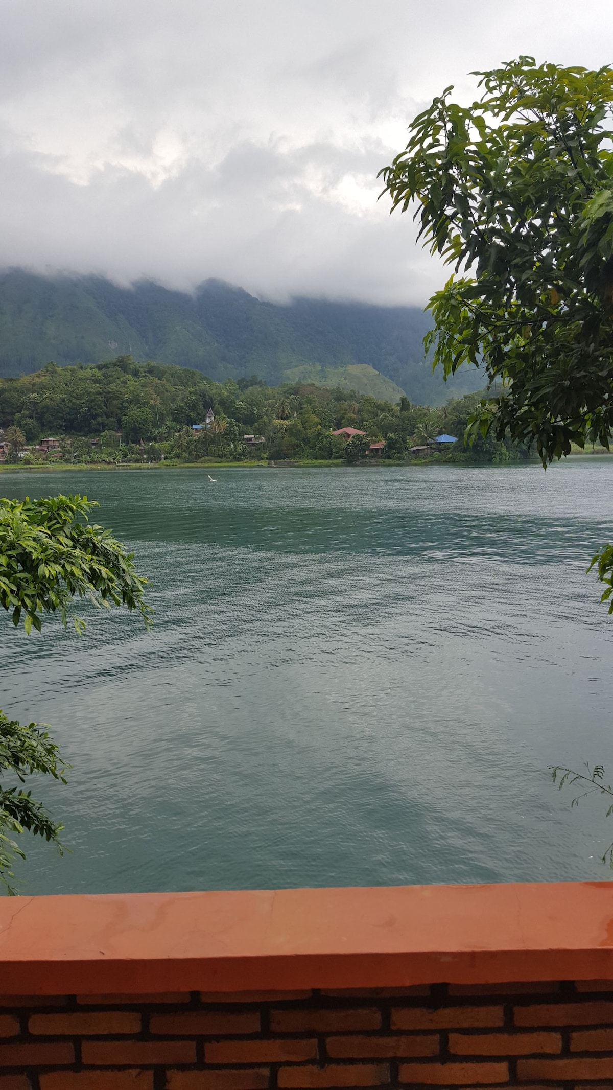The serene lake is framed by lush green foliage and distant mountains, partially shrouded in clouds. Gentle waves create a calming surface, reflecting the surrounding nature. A brick wall is visible in the foreground, anchoring the view of the tranquil water.