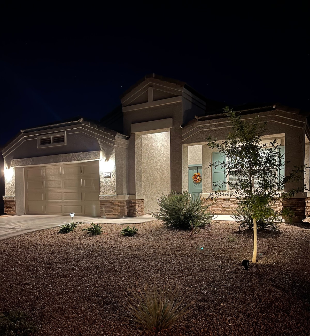 The exterior of the home is illuminated at night, highlighting modern architectural features and stone accents. Low-maintenance landscaping surrounds the entrance, which includes cheerful decorations and surrounded by desert plants. The garage door and entryway are well-defined in soft lighting.