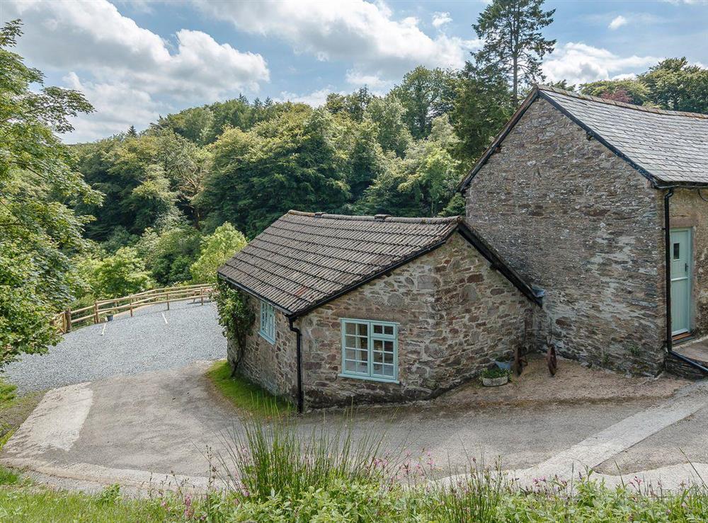 The exterior of the stone cottage is shown, set within a natural landscape surrounded by trees. A gravel driveway leads to the cottage, and a fenced area is visible in the background. The roof features a traditional design, complementing the tranquil setting.
