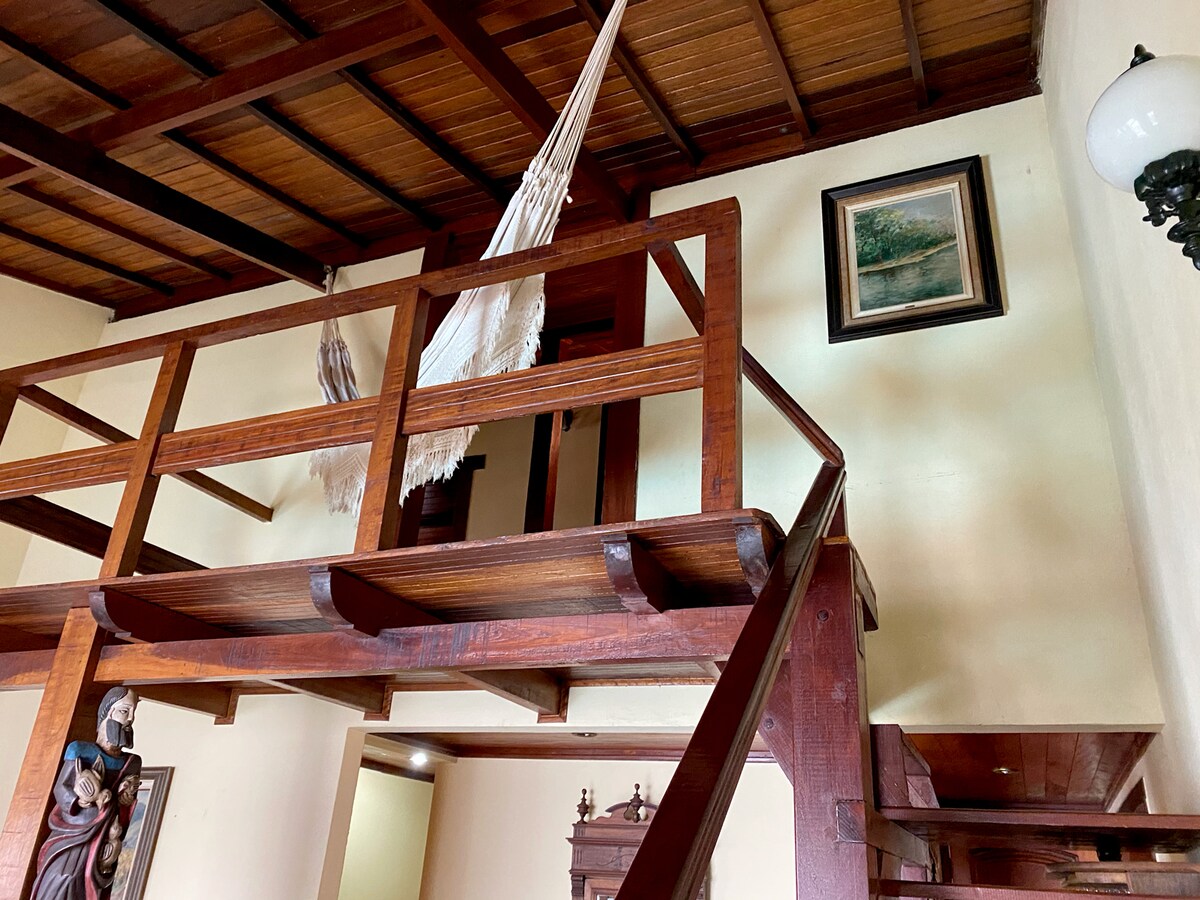 A wooden loft area is visible above a staircase, highlighted by a suspended hammock and artwork on the wall. The warm tones of the hardwood ceiling complement the natural elements of the space, creating a cozy and inviting ambiance.