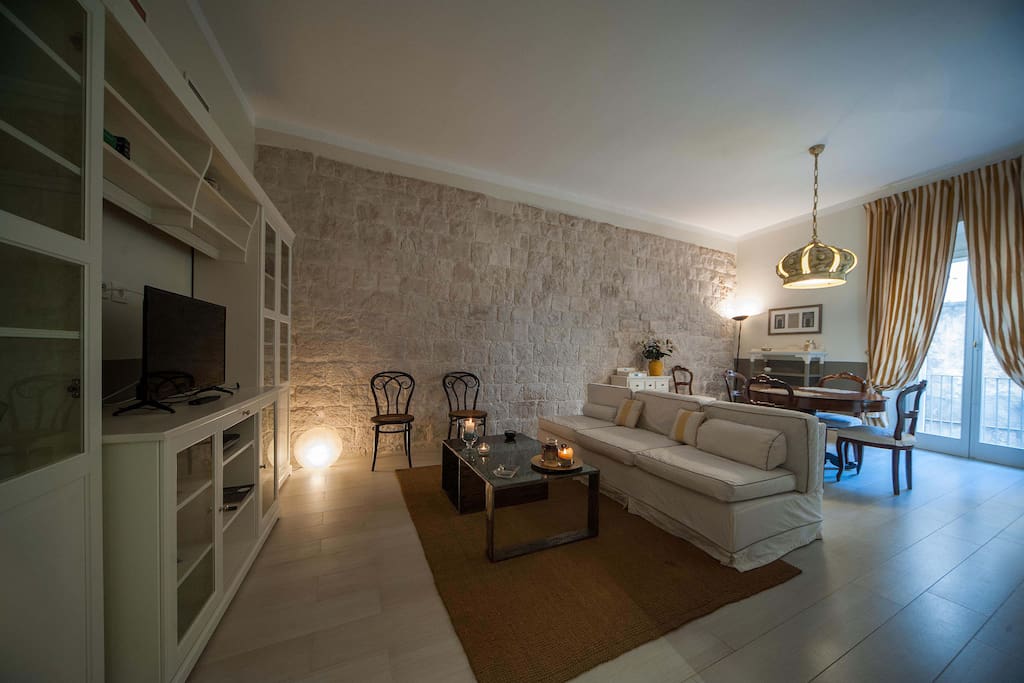 Zona leaving e pranzo: stanza ampia e luminosa con parete in pietra naturale e archi di passaggio illuminati; zona relax con divano, tv e parete attrezzata + zona con tavolo pranzo