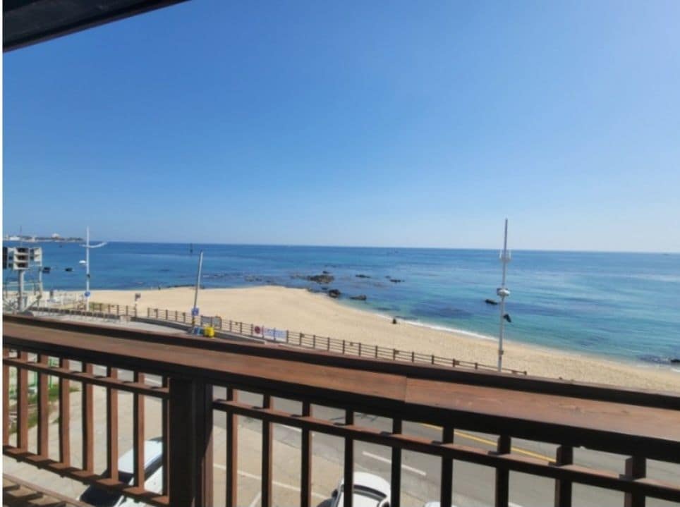 A beachfront view captures calm ocean waters and a sandy shore, framed by a wooden railing. Clear skies create a serene atmosphere, while distant boats float gently on the horizon.