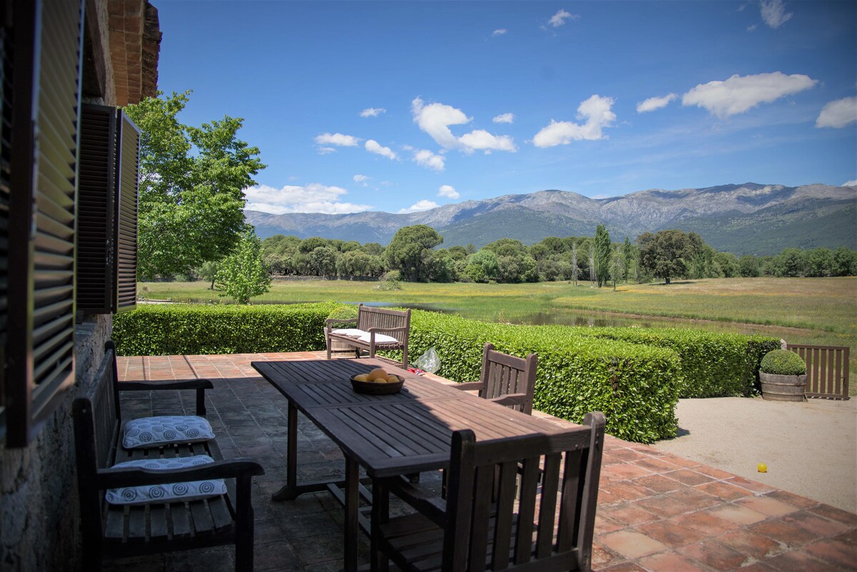 An outdoor terrace features a large wooden table surrounded by several matching chairs. The view includes rolling green fields and distant mountains under a clear blue sky, with trees framing the landscape. The area is well-maintained, offering a relaxing setting.
