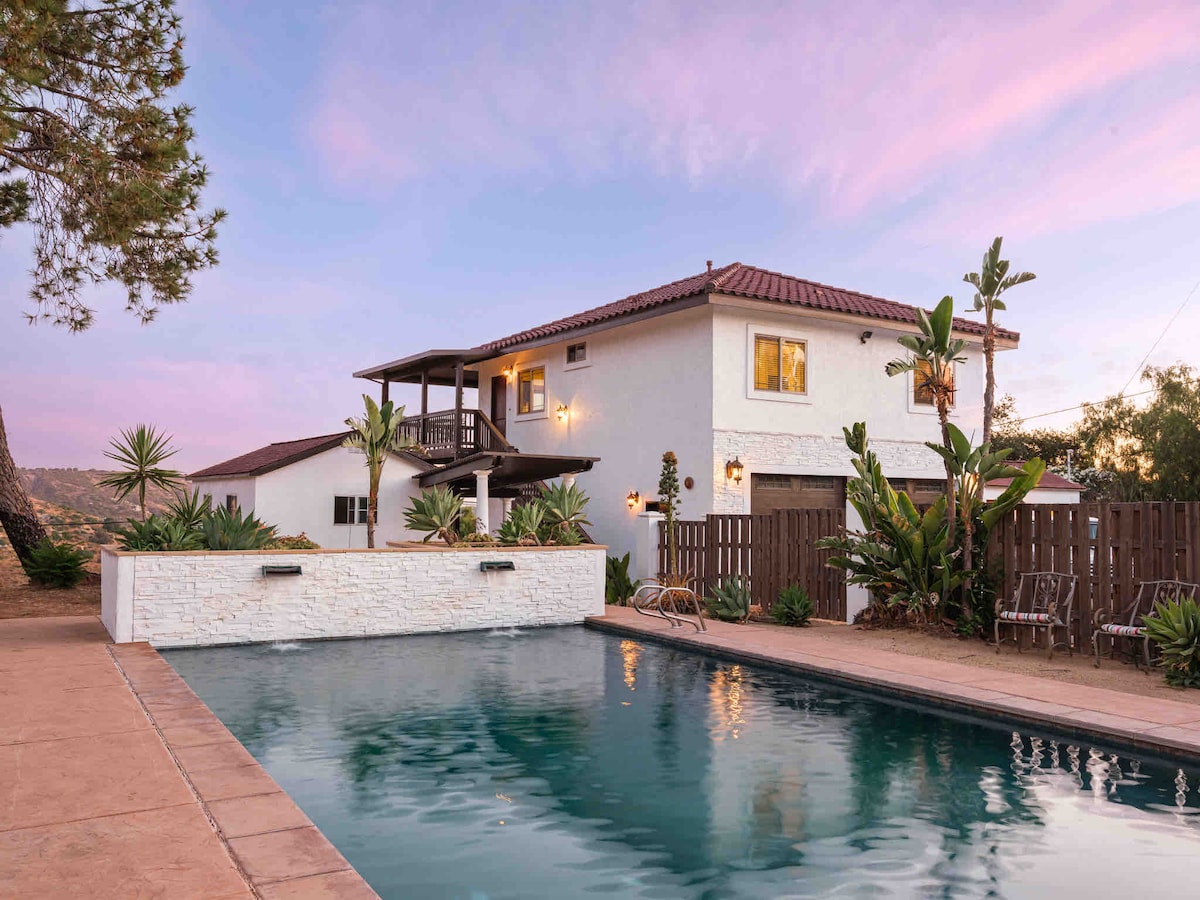A two-story guesthouse is pictured next to a tranquil pool, surrounded by lush greenery. The building features multiple balconies and large windows, offering views of the surrounding landscape. A stone wall runs alongside the pool area, enhancing the outdoor ambiance.