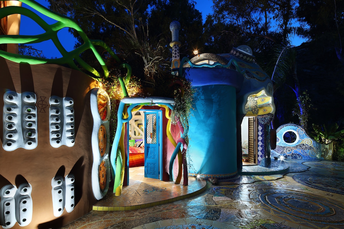 A unique architectural structure is illuminated at night, showcasing vibrant colors and intricate designs. Doorways lead into the whimsical interiors, surrounded by lush greenery and artistic features. A winding pathway accentuates the playful layout of the space, inviting exploration.