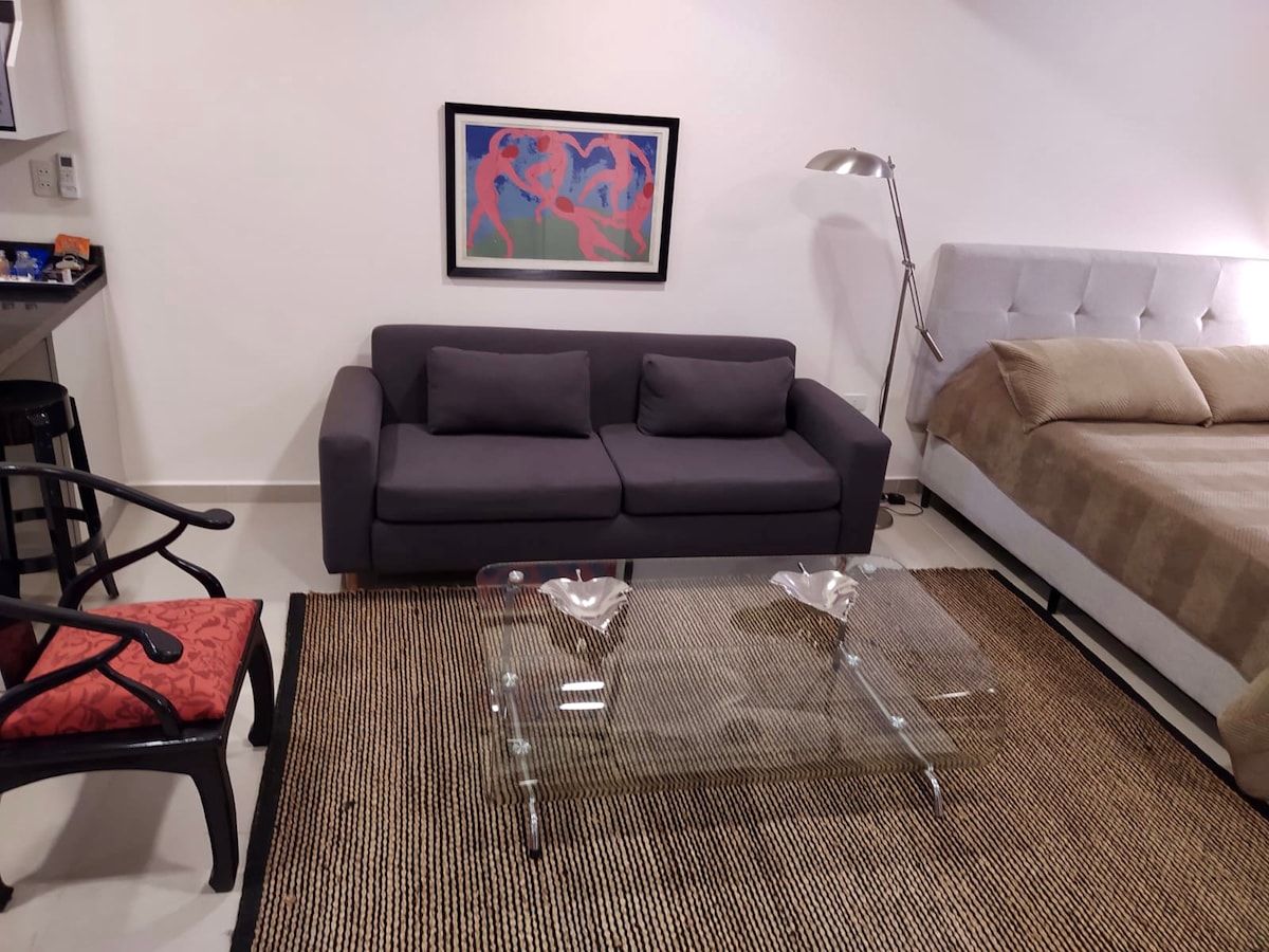 A cozy studio setting features a gray sofa with two cushions facing a glass coffee table. A spacious bed with a light-colored coverlet is positioned nearby. An artwork in bold colors is hung on the wall above the sofa, adding a touch of character to the room.