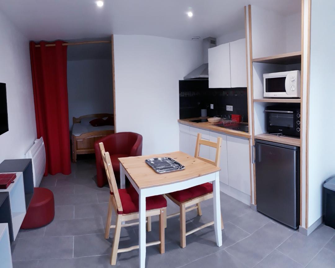 A living area features a wooden dining table surrounded by four chairs, with red cushions. A kitchenette, equipped with modern appliances, is visible. A small lounge area with two red chairs is present. Soft lighting illuminates the space, enhancing its functional layout.