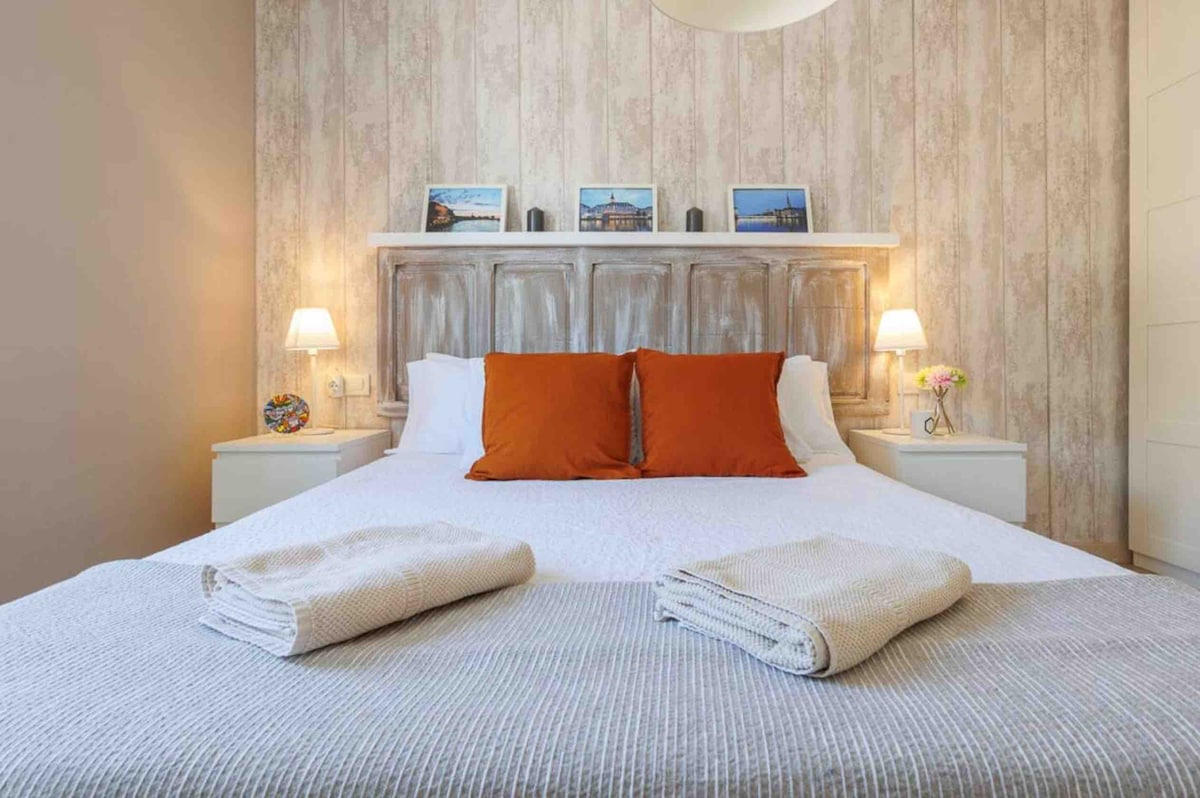 A spacious bedroom is highlighted by a wooden headboard adorned with framed photographs. Two bedside lamps provide soft lighting, complementing orange accent pillows on the large bed. Fresh towels are neatly placed at the foot of the bed, with a decorative floral arrangement present on the nightstand.