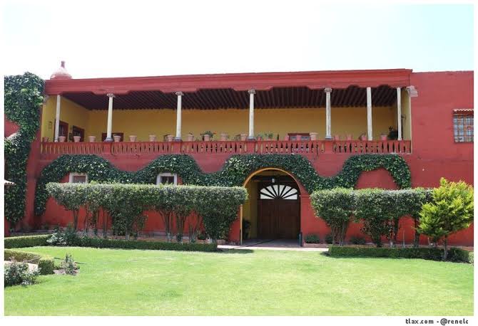 The exterior of the Ex-Hacienda San Buenaventura is showcased, featuring a vibrant red facade adorned with lush greenery. A covered balcony supported by columns extends across the front, while neatly trimmed hedges and a manicured lawn enhance the inviting appearance of the property.