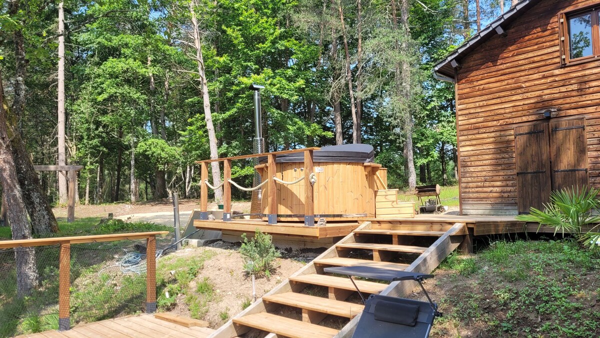 A wooden deck surrounds a Nordic bath positioned near a rustic chalet. The bath, designed for six, is complemented by natural greenery and trees in the background, creating a serene environment. Steps lead from the deck to the ground, enhancing accessibility.