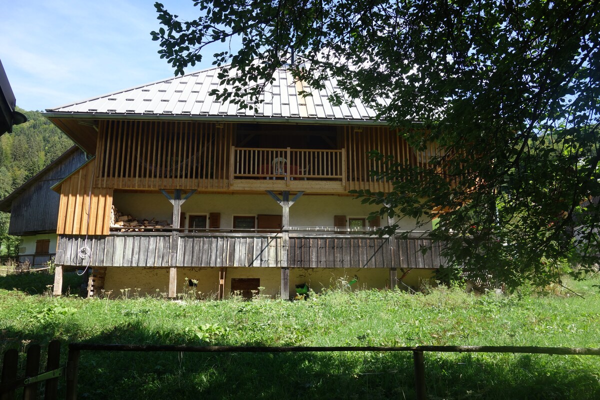 The exterior of the traditional farmhouse is framed by greenery, showcasing a wooden balcony on the upper level. The sloped roof features tiles, while the wooden structure blends harmoniously with the surrounding landscape, presenting an inviting outdoor space beneath tall trees.
