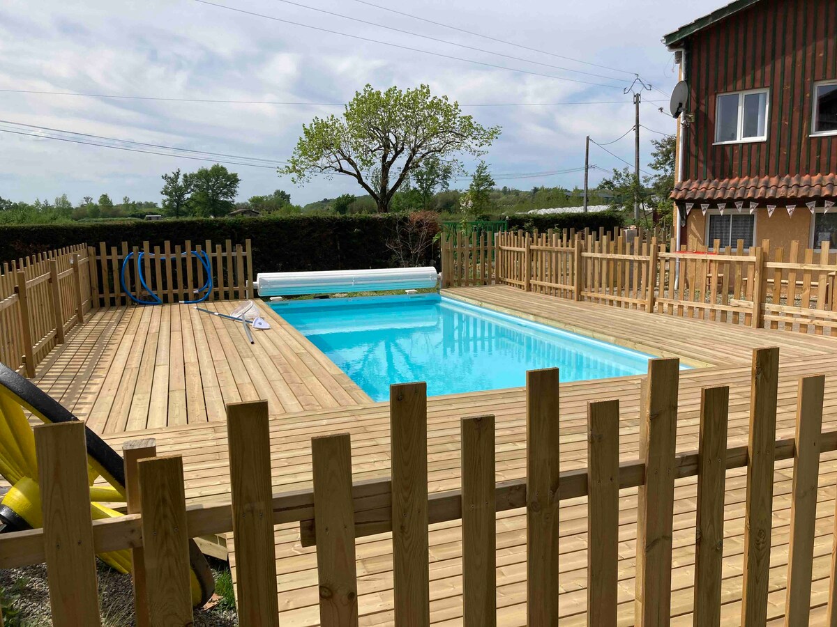 A wooden deck surrounds a clear swimming pool, inviting relaxation. The area is enclosed by a wooden fence, providing privacy. Green trees and shrubs are visible in the background, complementing the serene outdoor space.