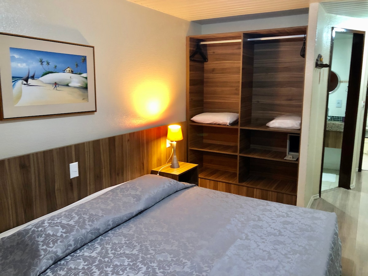 A neatly arranged sleeping area features a spacious bed covered with a grey patterned blanket. A bedside lamp emits a soft glow, while a framed artwork and wooden closet with shelves provide a warm touch. Light wooden accents enhance the overall ambiance.