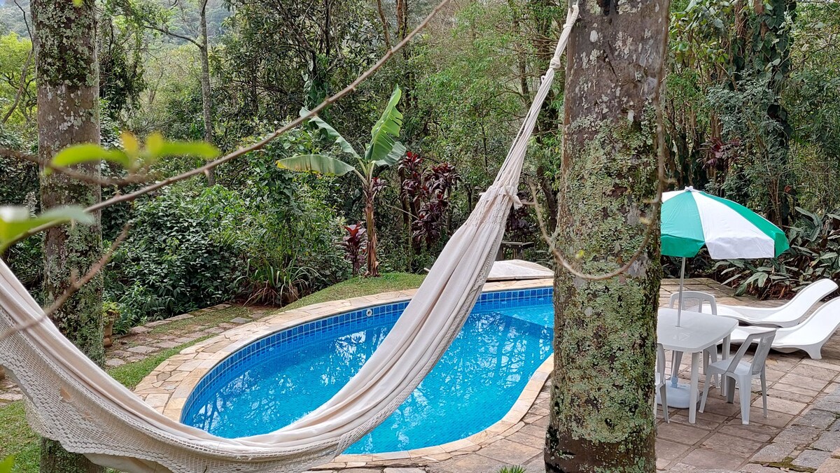 A hammock is suspended between two trees, providing a relaxing spot near a shimmering pool surrounded by lush green foliage. Lounge chairs and a table with an umbrella can be seen nearby, inviting guests to enjoy the tranquil outdoor space.